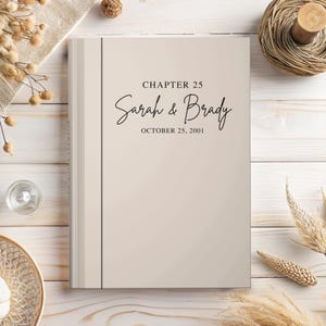 May include: A light beige hardcover book with a vertical dark gray line on the left side. The cover features the black printed text "CHAPTER 25", the names "Sarah & Brady" in a cursive script, and the date "OCTOBER 25, 2001" centered on the front.