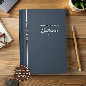 May include: A dark gray journal with the words "Word of the Year Balance" and a heart symbol. The journal has a white vertical stripe and the text "2026 Intentions". A pencil and a plant are also visible.