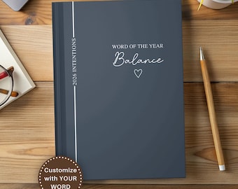 May include: A dark gray journal with the words "Word of the Year Balance" and a heart symbol. The journal has a white vertical stripe and the text "2026 Intentions". A pencil and a plant are also visible.