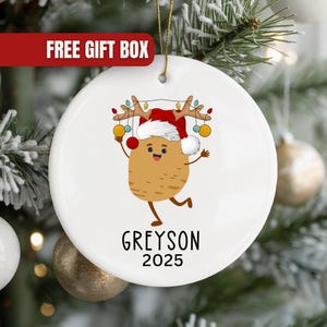 May include: White ceramic Christmas ornament featuring a cartoon potato character wearing a Santa hat and reindeer antlers, with colorful lights. The name "Greyson" and the year "2025" are printed below the character. The ornament hangs on a Christmas tree.