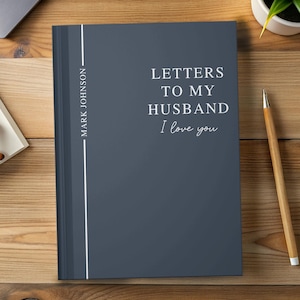 May include: A dark blue journal with the text "LETTERS TO MY HUSBAND I love you" in white. The spine has the name "MARK JOHNSON" in white. A pencil and a small plant are on the wooden surface.