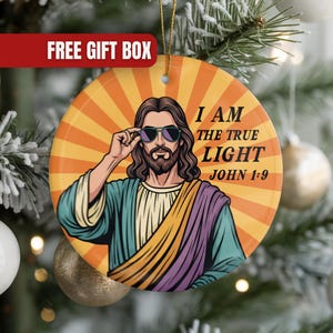 May include: Round Christmas ornament featuring an illustration of Jesus wearing sunglasses, set against a retro orange and yellow burst design. The text reads "I AM THE TRUE LIGHT JOHN 1:9". The ornament is hanging on a Christmas tree.
