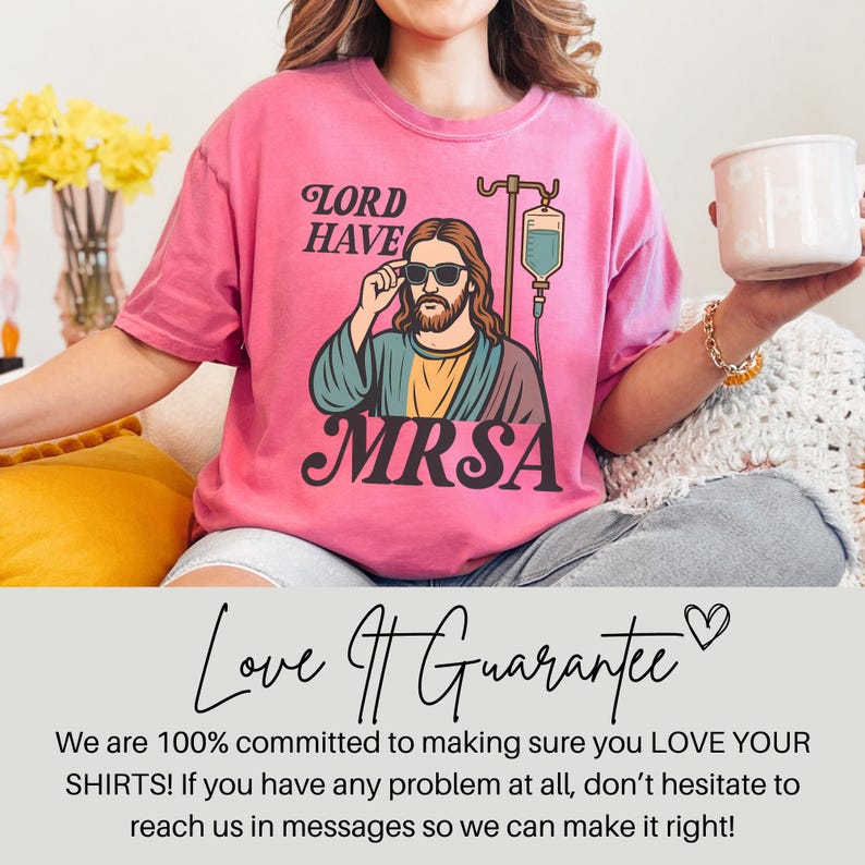 May include: Pink t-shirt with a graphic of Jesus wearing sunglasses and an IV drip, with the text "LORD HAVE MRSA". The shirt is a vibrant pink color and has a relaxed fit. The design is a humorous take on a religious theme.