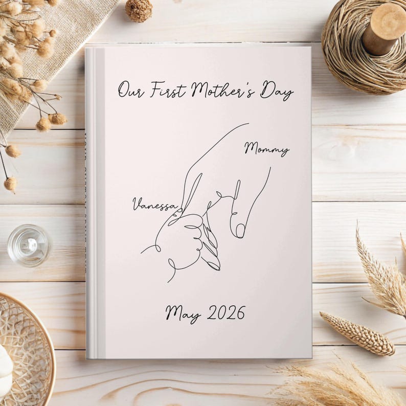 May include: A pale pink journal featuring the text "Our First Mother's Day" and a line drawing of a hand holding a baby's hand. The names "Vanessa" and "Mommy" are included, along with the date "May 2026".