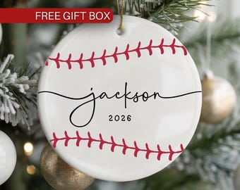 May include: A round white ceramic ornament featuring red baseball stitching graphics across the top and bottom. The centre displays the name Jackson in black cursive script with the year 2026 printed below in a simple black sans-serif font.
