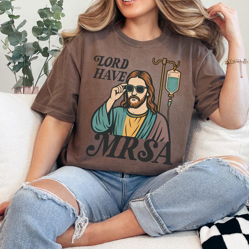 May include: Brown t-shirt with a graphic of Jesus wearing sunglasses and an IV drip, with the text "LORD HAVE MRSA". The graphic features Jesus in a colorful robe. The shirt is worn by a person wearing blue jeans.