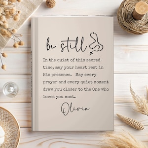 May include: A beige journal with the words "be still" in black script. The journal includes a rosary and cross illustration, and the text "In the quiet of this sacred time...Olivia."