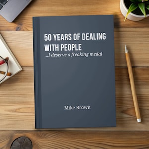 May include: A hardback book with a dark blue cover titled "50 Years of Dealing With People" with the subtitle "...I deserve a freaking medal" and the author's name, Mike Brown. The book is on a wooden surface with a pencil and a small plant.
