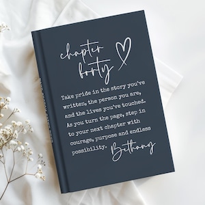 May include: A navy blue journal with the words "chapter forty" and a heart symbol in white script. The journal includes inspirational text and the name "Bethany" in cursive. The journal is on a white surface.