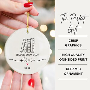 May include: A round white ceramic ornament featuring a black line drawing of four books, the text "WILLOW BOOK CLUB", the name "olivia" in script, a small red heart, and the year "2026". A gold string is attached to the top for hanging.