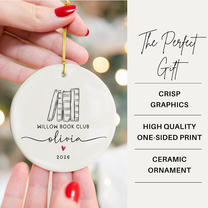May include: A round white ceramic ornament featuring a black line drawing of four books, the text "WILLOW BOOK CLUB", the name "olivia" in script, a small red heart, and the year "2026". A gold string is attached to the top for hanging.