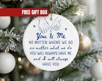 May include: A white ceramic Christmas ornament with a gold string, featuring blue text and a hand-holding illustration. The text reads "You & Me" and a loving message. The ornament hangs on a Christmas tree with white and gold ornaments.