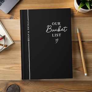 Pu&ograve; includere: Un diario nero con le parole "OUR Bucket List" in corsivo bianco, un piccolo cuore e i nomi "BRIAN & NICOLE" verticalmente sul dorso. Una matita di legno &egrave; appoggiata su una superficie di legno.