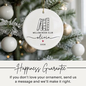 May include: A round white ceramic ornament featuring a black line drawing of a row of books, the text WILLOW BOOK CLUB, the name olivia in script, a small red heart, and the year 2026. The ornament hangs from a gold string against a tree.