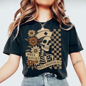 May include: Black t-shirt featuring a skeleton in a nurse's hat with a cross, surrounded by flowers. The shirt has the text "Sorta Ratchet Sorta Nightingale" on a banner. A checkered pattern is on the right side of the design.