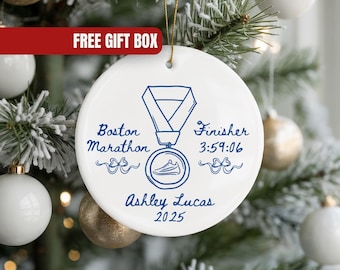 May include: White ceramic Christmas ornament with a blue illustration of a marathon medal. The ornament has the text "Boston Marathon Finisher 3:59:06 Ashley Lucas 2025". A red banner at the top reads "FREE GIFT BOX".