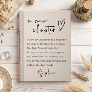 May include: A beige hardcover journal featuring the black text 'a new chapter' with a heart symbol, followed by a printed message about baptism and dedication to Jehovah, ending with the name 'Sophia' in a cursive script font on the cover.