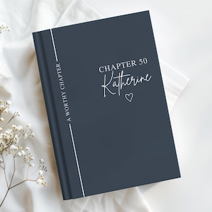 May include: A navy blue hardcover journal with the text "Chapter 50 Katherine" in white script, with a small heart below the name. The spine features a vertical white line and the words "A Worthy Chapter."