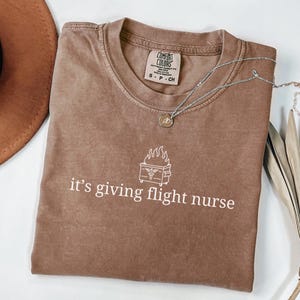May include: A brown t-shirt with a white graphic of a flaming dumpster with a medical symbol inside. The text "it's giving flight nurse" is printed below the graphic.