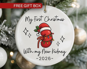 May include: A round white ceramic ornament featuring a red kidney illustration wearing a Santa hat and wrapped in colorful Christmas lights. The text reads My First Christmas With my New Kidney -2026- with small black star icons.