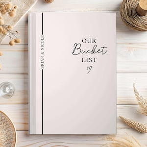 Pu&ograve; includere: Un diario di carta bianca con la scritta "OUR Bucket LIST" in corsivo e un simbolo a forma di cuore. Sul dorso sono riportati i nomi "BRIAN & NICOLE". Il diario &egrave; su una superficie di legno chiaro.
