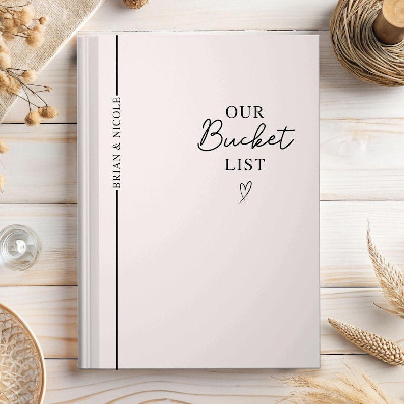 Pu&ograve; includere: Un diario di carta bianca con la scritta "OUR Bucket LIST" in corsivo e un simbolo a forma di cuore. Sul dorso sono riportati i nomi "BRIAN & NICOLE". Il diario &egrave; su una superficie di legno chiaro.