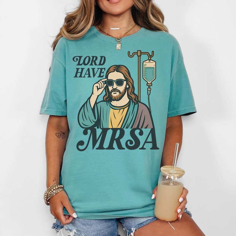 May include: Teal-colored t-shirt with a graphic of Jesus wearing sunglasses, with the words "LORD HAVE" above and "MRSA" below. The graphic includes an IV drip bag. The shirt is a casual, unisex style.