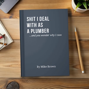 May include: A dark blue book titled "SHIT I DEAL WITH AS A PLUMBER" with the subtitle "...and you wonder why I cuss" by Mike Brown. The book is on a wooden surface with a pencil, a small plant, and a laptop visible.