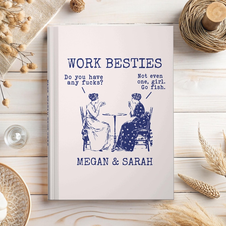 May include: A white book cover featuring blue text and a vintage-style illustration of two people sitting at a table playing cards. The text reads: WORK BESTIES, Do you have any fucks?, Not even one, girl. Go fish., and MEGAN & SARAH.