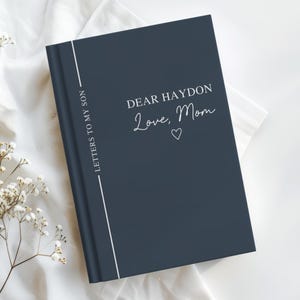 May include: A navy blue journal with the text "DEAR HAYDON Love, Mom" in white script. The journal has a white vertical line on the left side and the words "LETTERS TO MY SON" printed vertically. A small white heart is below "Mom".