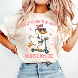 May include: Cream-colored t-shirt featuring a cartoon duck wearing sunglasses and a bow, riding a scooter. The text "HELP IS ON THE WAY" and "NURSE KYLEE" are printed in pink. Pink stars surround the duck.