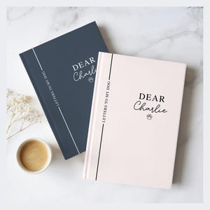 May include: Two hardcover journals, one navy blue and one light pink, with the text "DEAR Charlie" and a paw print. The spine reads "LETTERS TO MY DOG". A small cup of coffee and sprigs of white flowers are in the background.