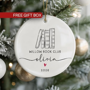 May include: A round white ceramic ornament featuring a black line drawing of a stack of books. The text reads WILLOW BOOK CLUB, the name olivia in script, and the year 2026, with a small red heart icon centered below the name and a gold string.