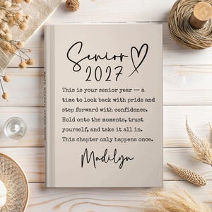 Op de afbeelding: Een beige notitieboek met de woorden "Senior 2027" en een hartsymbool in zwart schrift. Het notitieboek bevat tekst over het laatste jaar en de naam "Madilyn". Het notitieboek ligt op een licht houten oppervlak.