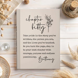 May include: A hardcover journal with the words "chapter fifty" in elegant script, accompanied by floral illustrations. The journal includes inspirational text and the name "Bethany" at the bottom, set against a neutral background.