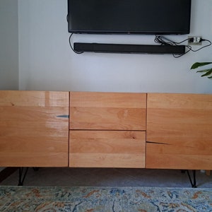 May include: A light wood entertainment center with three drawers and two doors. The cabinet has black metal hairpin legs and is sitting on a patterned rug.