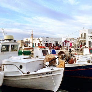Pode incluir: Um grupo de barcos de pesca brancos e azuis atracados num porto. Os barcos estão amarrados juntos e têm vários nomes e números pintados nos seus lados, incluindo "DANAIITA MI 156" e "IANKETIS 254". Os barcos estão em frente a uma fila de edifícios brancos com portas e janelas vermelhas.