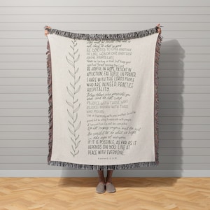 May include: A cream-coloured throw blanket with a black and white text design. The text is a bible verse from Romans 12:9-18. The blanket has a fringe border and a subtle floral pattern.