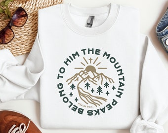 Könnte beinhalten: Weißes Sweatshirt mit einem kreisförmigen Grafikdesign mit einer Bergkette, Bäumen und dem Text "BELONG TO HIM THE MOUNTAIN PEAKS" und "PSALM 95:4" in Grün und Braun. Das Sweatshirt ist auf einer weißen Oberfläche gefaltet.