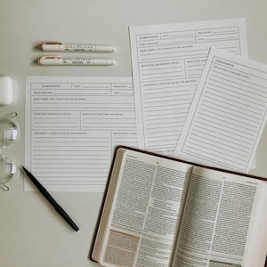 Può includere: Una vista dall'alto di una scrivania bianca con una penna nera, due evidenziatori bianchi, un bicchiere trasparente, una custodia bianca per auricolari wireless, una Bibbia rilegata in pelle marrone e due fogli di carta bianchi con la scritta "Sermon Notes" stampata su di essi.