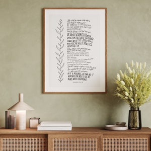 Può includere: Una stampa incorniciata con una scrittura a mano di un versetto biblico, con un design di foglie decorative. La stampa è in una cornice di legno ed è esposta su un mobile di legno. Il testo include le parole "Vivete in pace con tutti."
