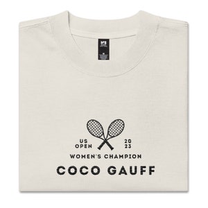 May include: A white t-shirt with a black graphic of two tennis rackets crossed over each other. The text "US OPEN 2023 WOMEN'S CHAMPION COCO GAUFF" is printed below the rackets.