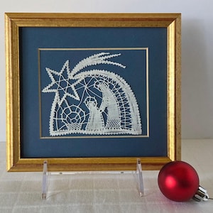 May include: A framed, white lace nativity scene with a gold frame. The artwork depicts a star, Mary, and baby Jesus on a dark blue background. A red Christmas ornament sits in front of the frame.