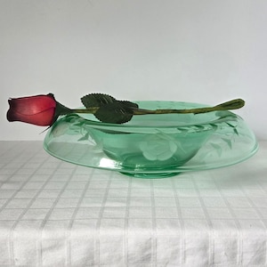 May include: A green glass bowl with etched floral designs, holding a red rose. The bowl has a wide, shallow shape and a pedestal base. The rose has a dark red bloom and green leaves.
