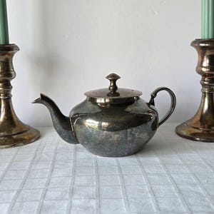May include: A vintage silver teapot with a curved spout, a rounded body, and a small handle. The teapot has a lid with a small knob on top. The teapot is placed on a white checkered tablecloth. Two brass candlesticks with green candles are on either side.