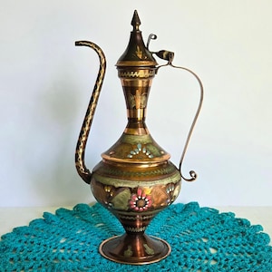 May include: A tall, slender metal ewer with a long, curved spout and a matching handle. The body features a polished copper finish with intricate hand-etched patterns, accented by painted floral motifs in shades of pink, green, blue, and white.
