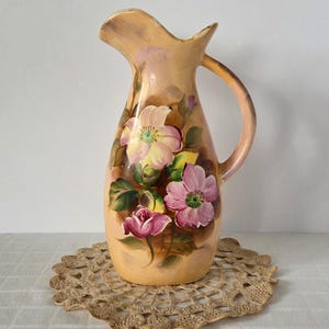 May include: A vintage ceramic pitcher with a peach-colored base and a curved handle. The pitcher is adorned with hand-painted pink and yellow flowers, green leaves, and rose buds. The pitcher is sitting on a beige lace doily.