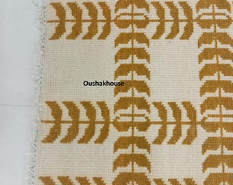 以下が含まれることがあります： 茶色の葉の幾何学模様が施された白い敷物。敷物はフリンジの縁取りがあり、敷物には「Oushakhouse」という文字が3回印刷されています。