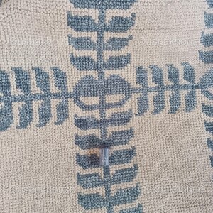 Op de afbeelding: Een klein, zilverkleurig metalen voorwerp met de letter "K" erop gegraveerd, liggend op een beige en blauw gepatroneerd tapijt.