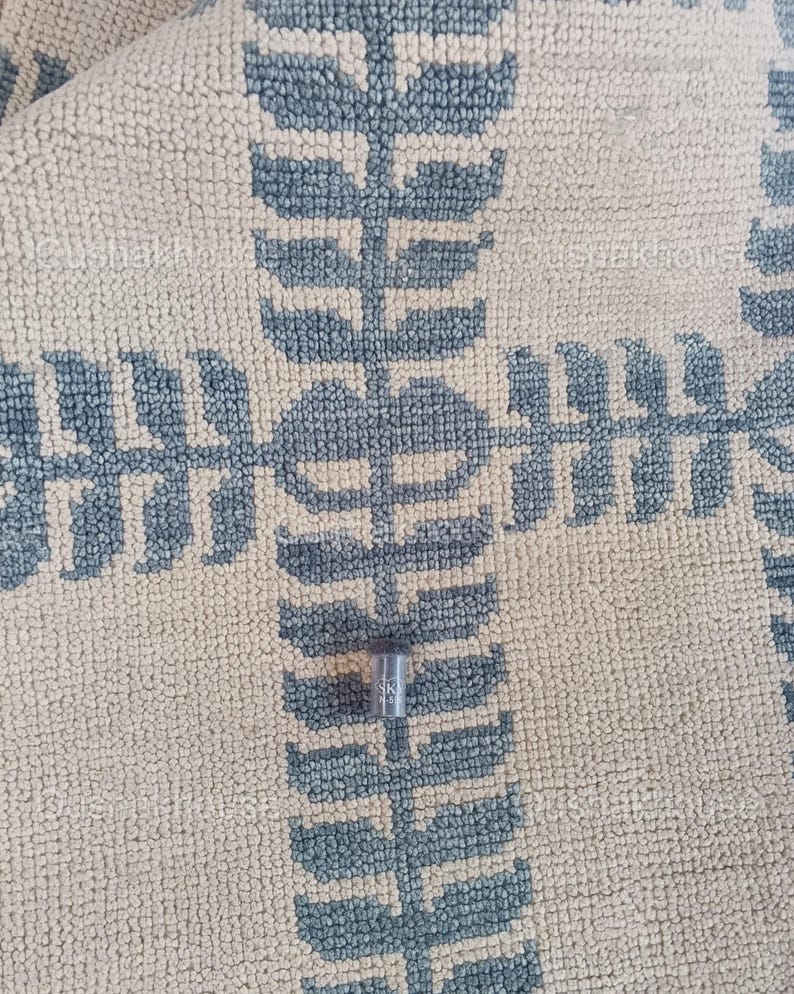 Op de afbeelding: Een klein, zilverkleurig metalen voorwerp met de letter "K" erop gegraveerd, liggend op een beige en blauw gepatroneerd tapijt.